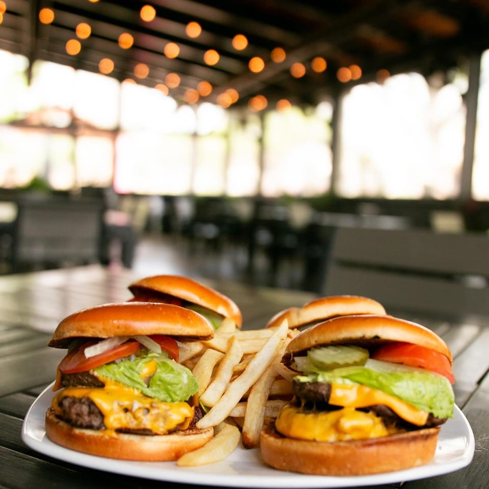 Two cheeseburgers with fries on a white plate, with a blurred restaurant patio in the background.