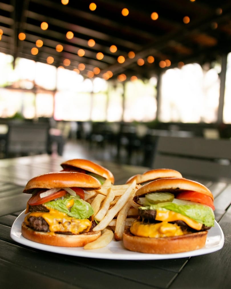 Two cheeseburgers with fries on a white plate, with a blurred restaurant patio in the background.