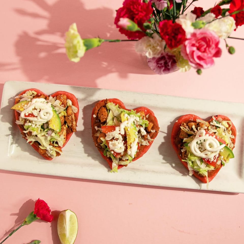 Three heart-shaped sopes with chicken, lettuce, avocado, and crema, on a white platter with flowers.