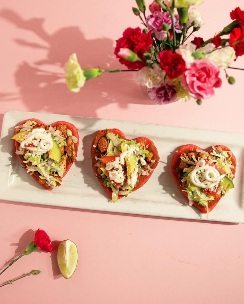 Three heart-shaped sopes with chicken, lettuce, avocado, and crema, on a white platter with flowers.