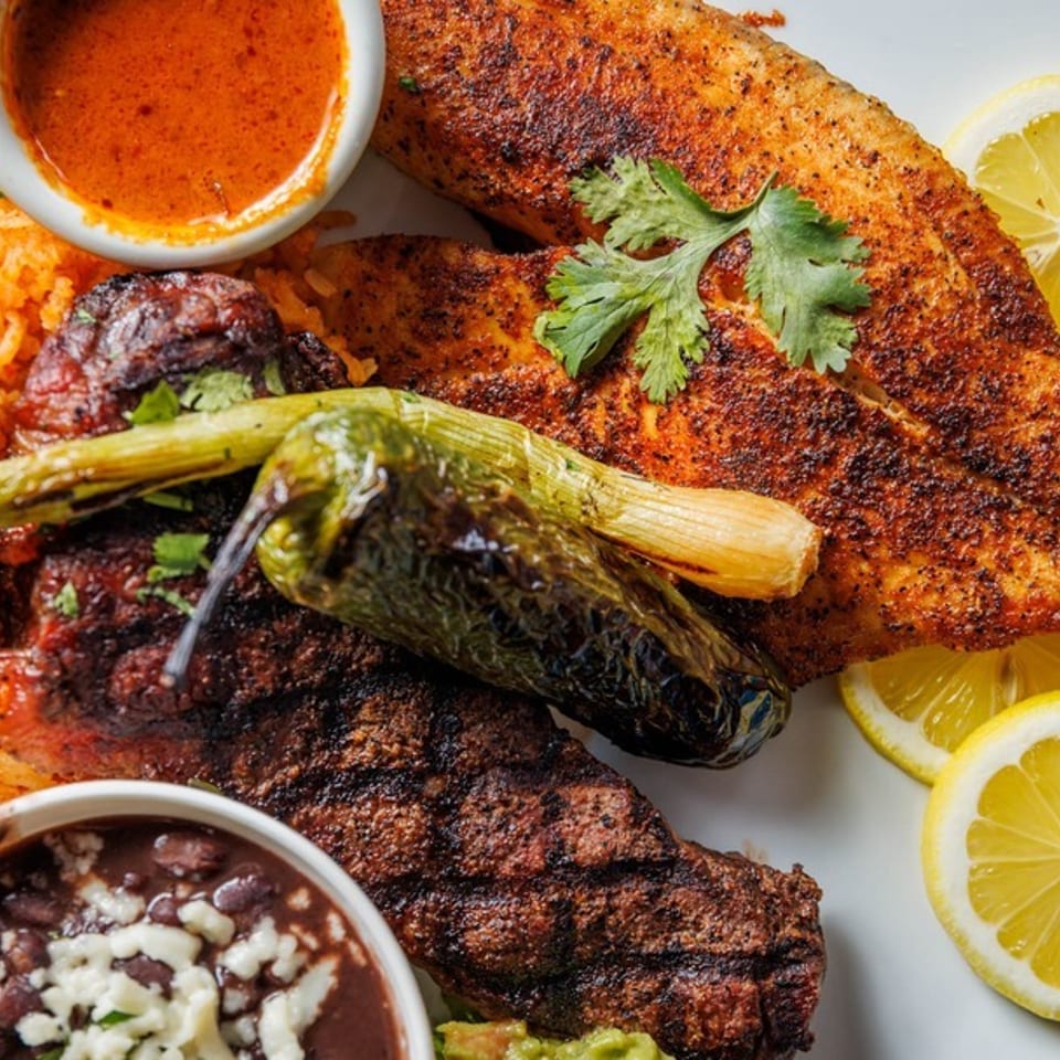 Grilled fish and steak with rice, beans, peppers, and lemon slices on a white plate.
