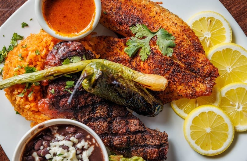 Grilled fish and steak with rice, beans, peppers, and lemon slices on a white plate.